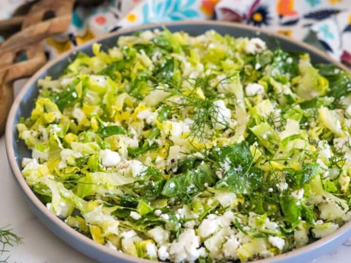 A bowl of chopped salad containing green leafy vegetables and crumbled white cheese, placed on a white marble surface with a colorful patterned cloth and wooden utensils in the background.