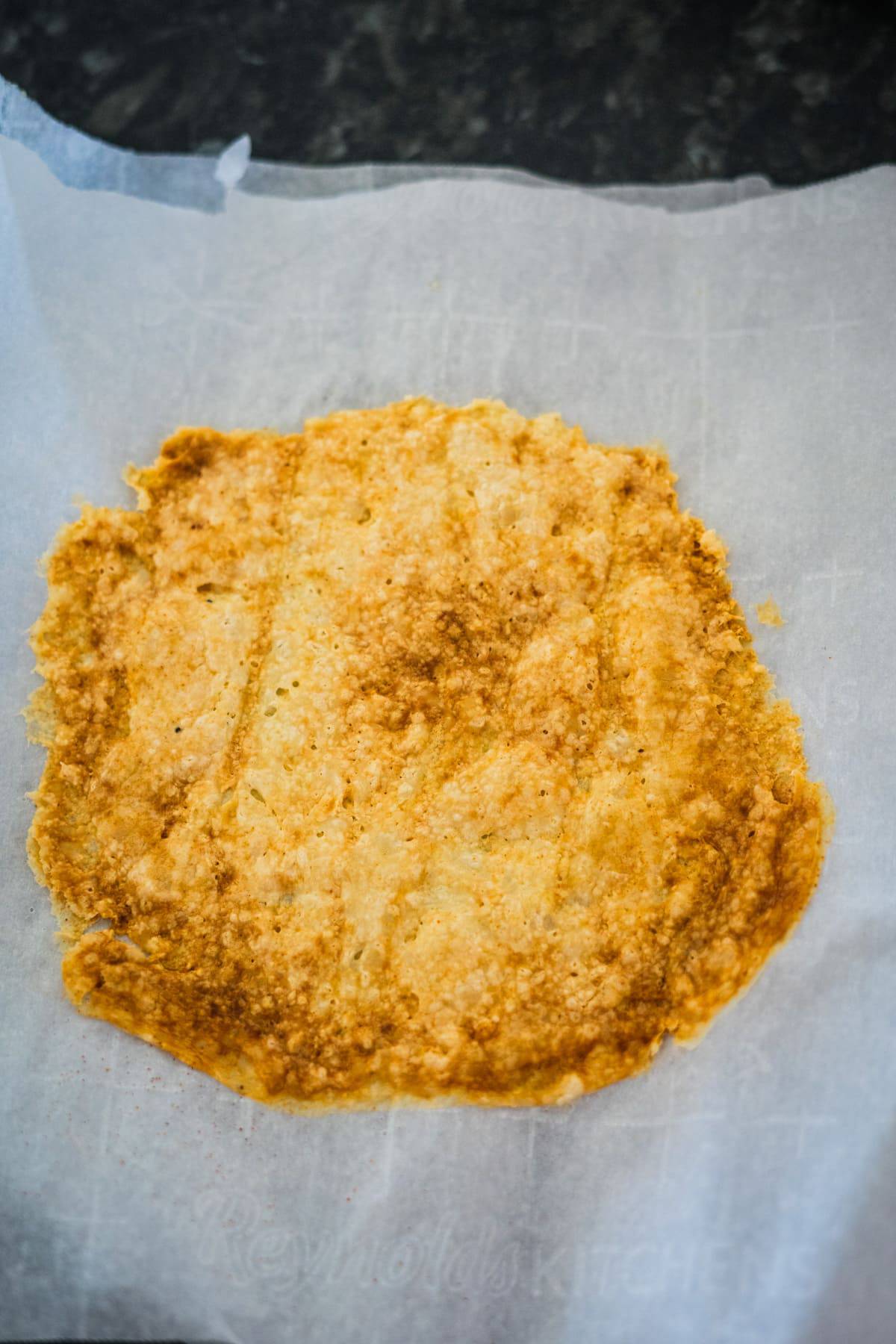 A baked, golden-brown flatbread or crust on a piece of parchment paper resting on a dark surface.