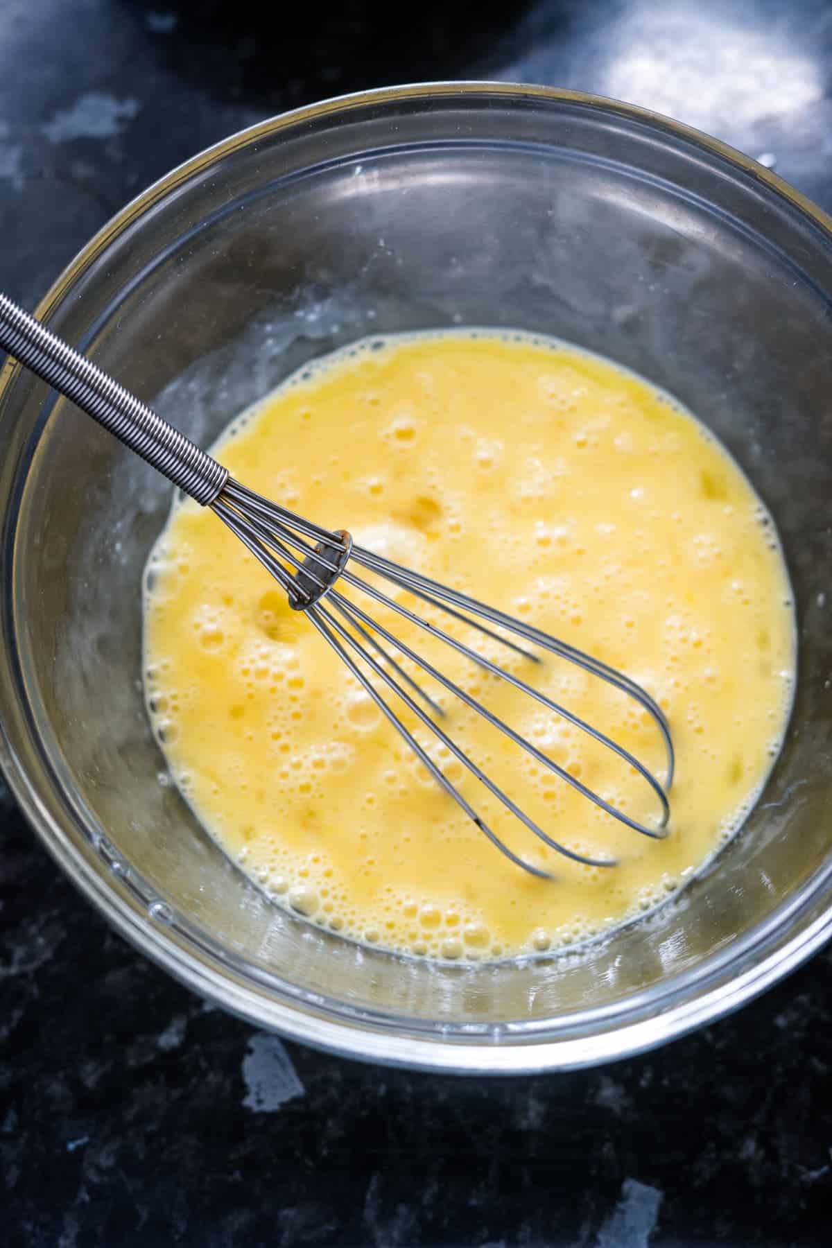 A whisk resting in a glass bowl containing beaten eggs on a dark countertop, ready to create delightful cottage cheese egg muffins.