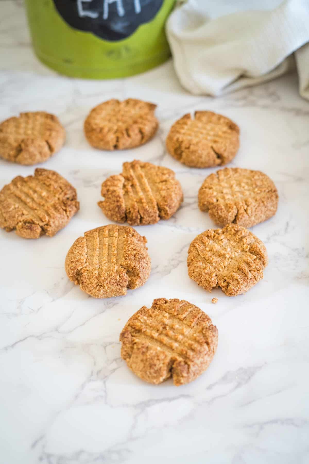 A batch of eight homemade peanut butter cookies on a white marble surface, with a green container and beige cloth in the background.