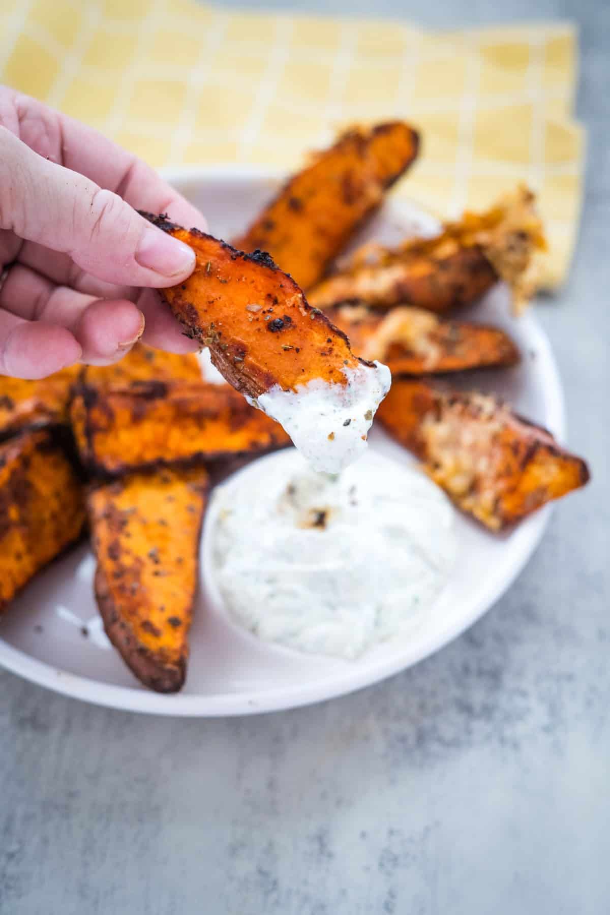 A hand dips a seasoned air fryer sweet potato wedge into a bowl of white dipping sauce, with more wedges on a plate in the background and a yellow napkin underneath.