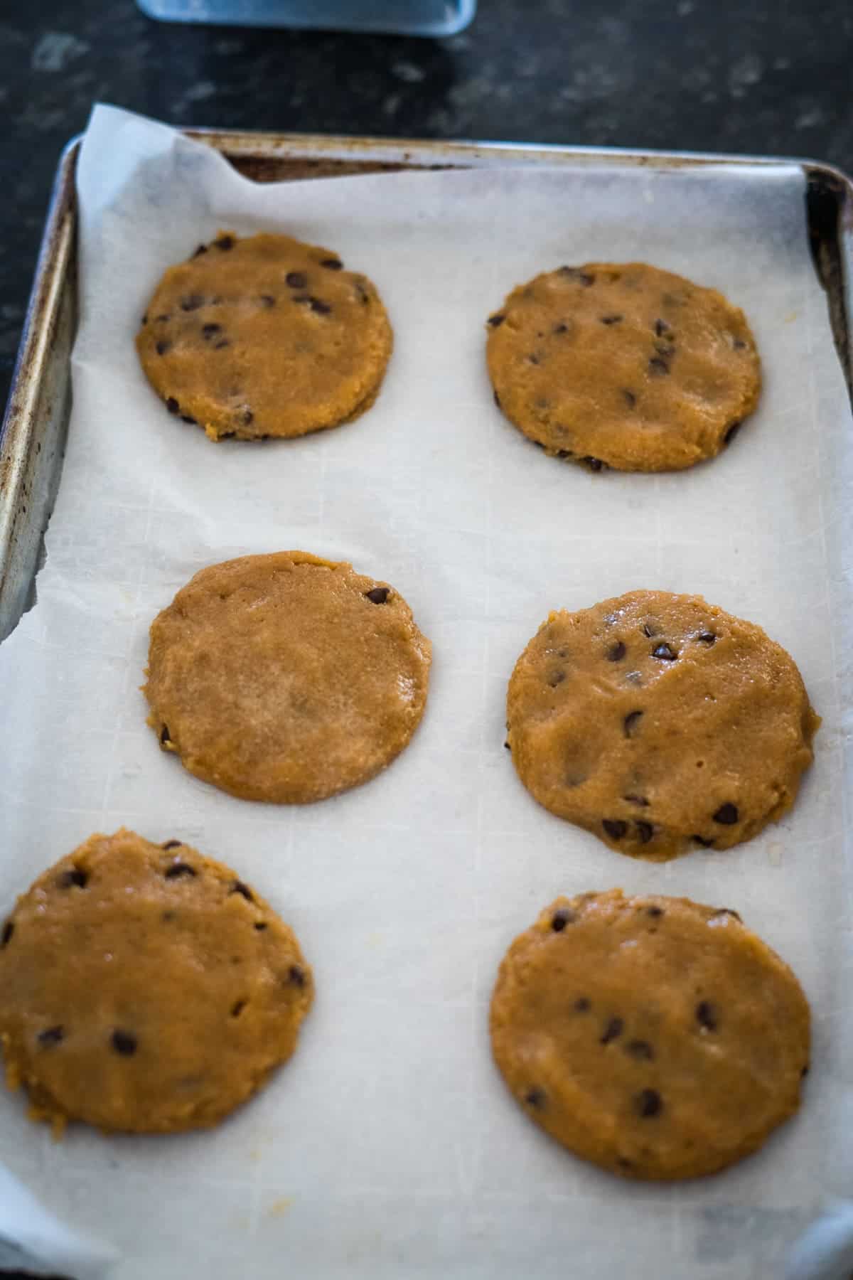 A baking sheet lined with parchment paper holding six evenly spaced, unbaked chocolate chip cookies.