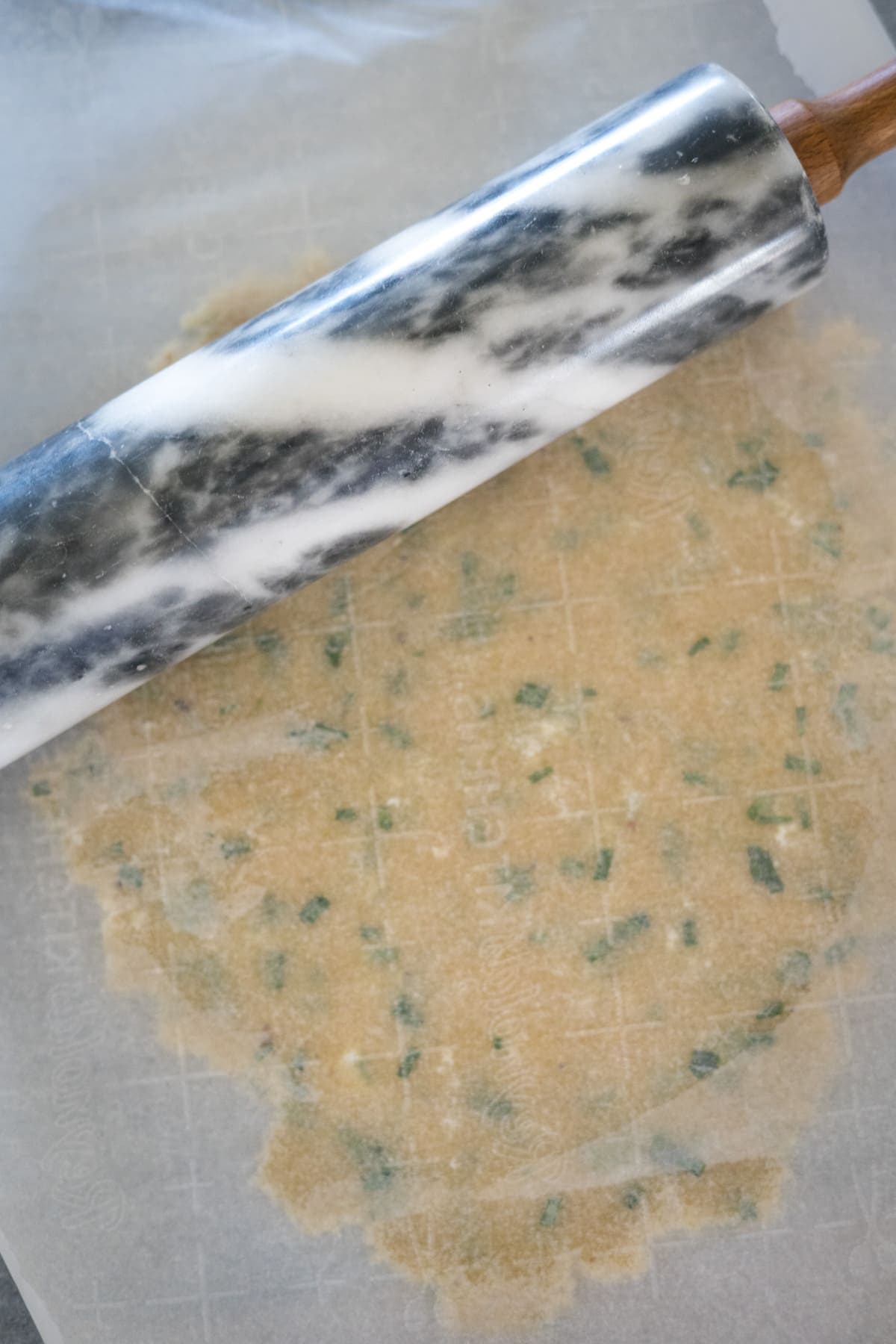 A marble rolling pin rolls out a sheet of dough with green herbs, placed on parchment paper.