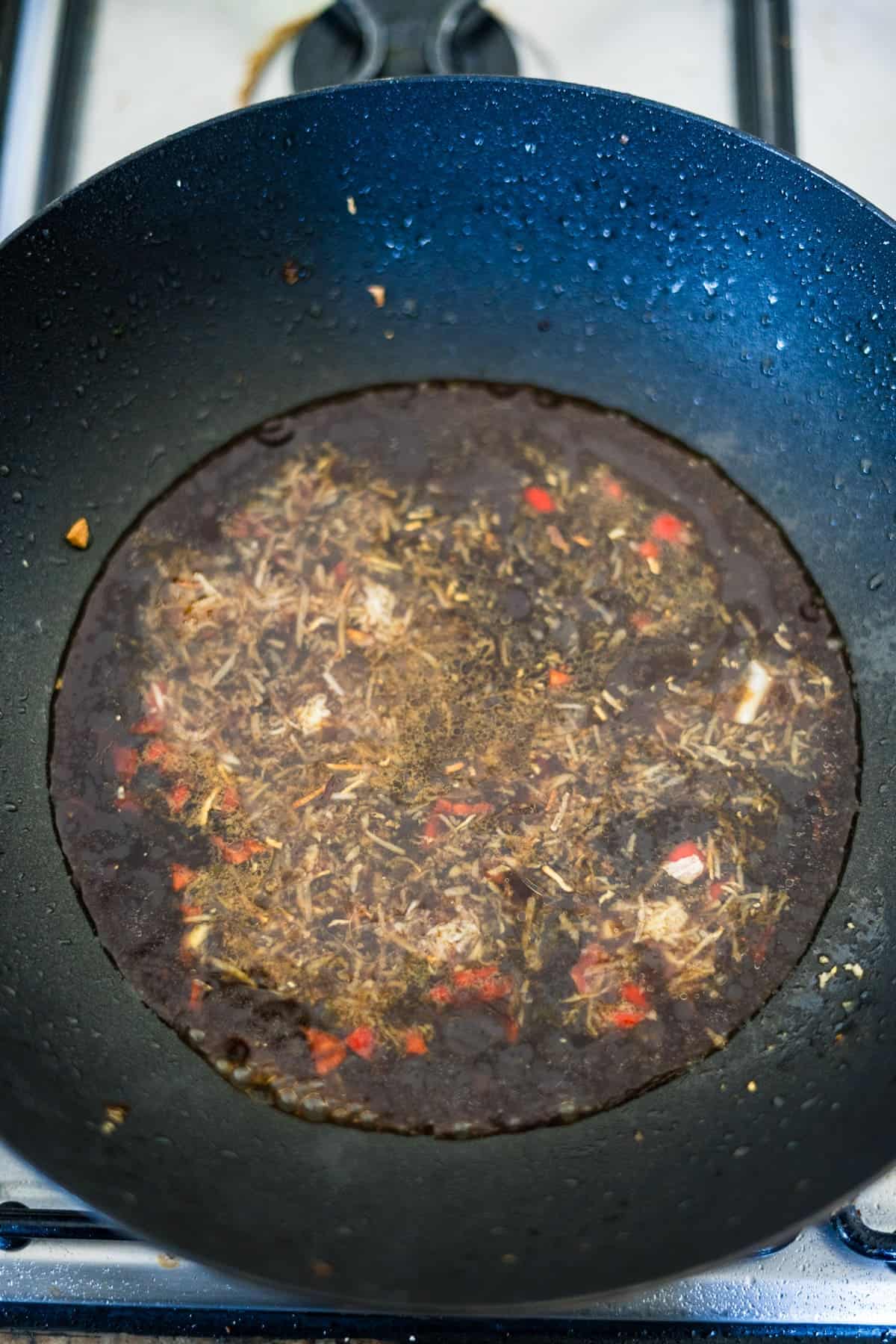 A black frying pan on a stovetop contains a rich, dark-colored liquid infused with floating herbs, spices, and small chunks of red and white ingredients from an eggplant tofu recipe.