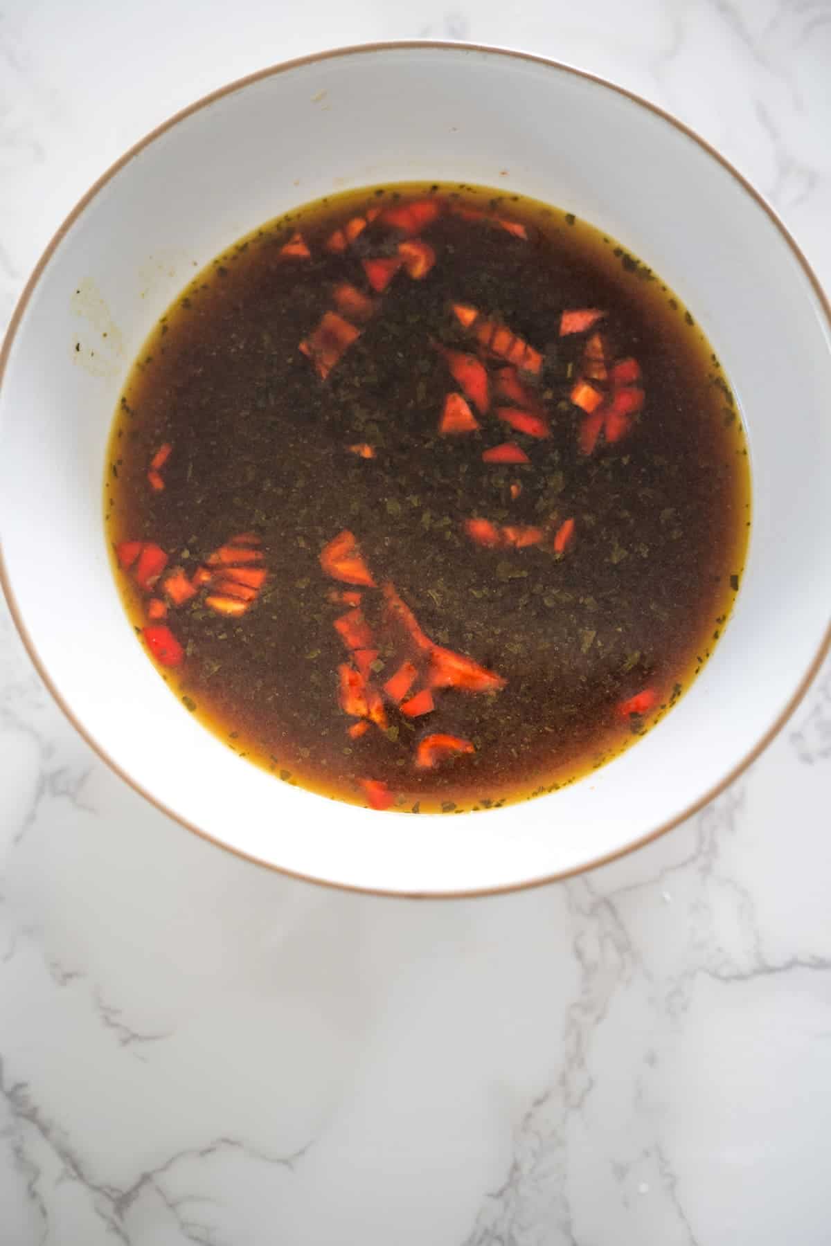 A white bowl filled with a dark, brownish-red broth containing pieces of red and orange vegetables, alongside tender eggplant, is placed on a white marble surface.