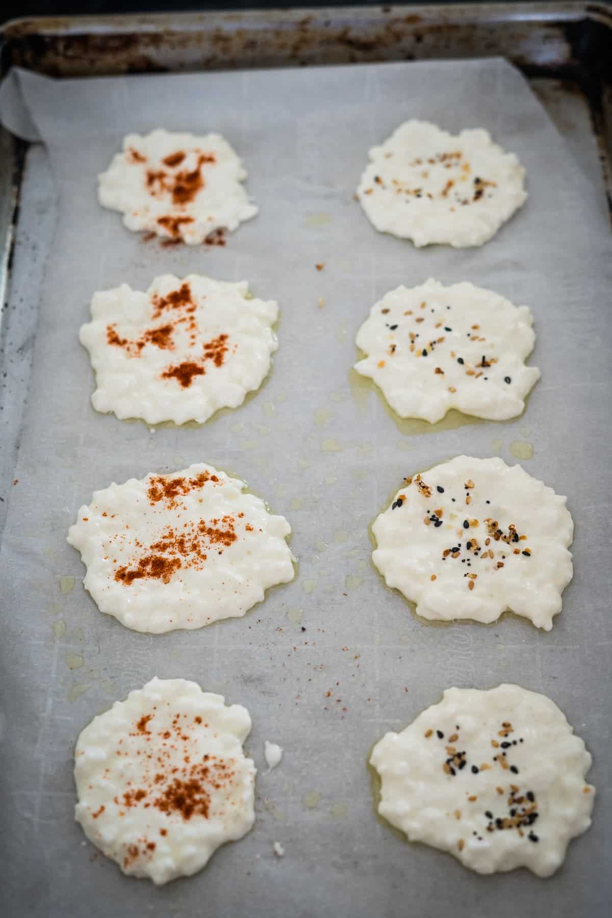 Eight uncooked rice cakes topped with various seasonings on a parchment-lined baking sheet.