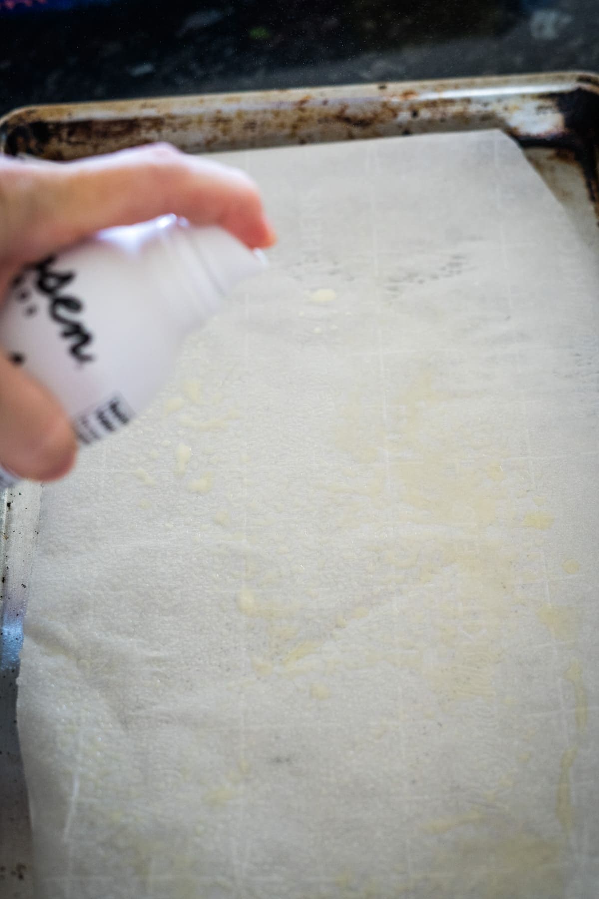 A person is spraying a can of oil onto a baking tray lined with parchment paper.