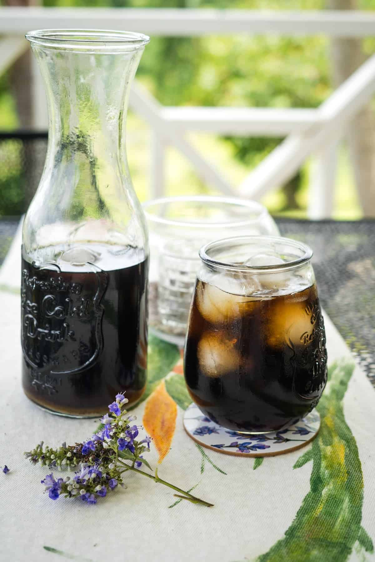 A glass pitcher and a small glass, both filled with dark sugar-free iced tea, are set on a floral tablecloth next to two empty glasses and purple flowers.