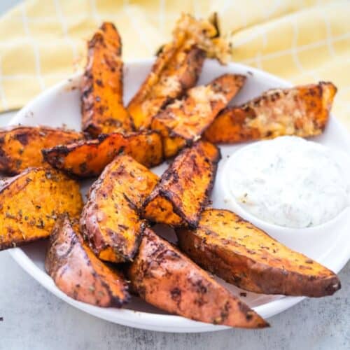 A white plate of seasoned sweet potato wedges with a side of white dipping sauce, placed on a light surface with a yellow-checkered cloth in the background.