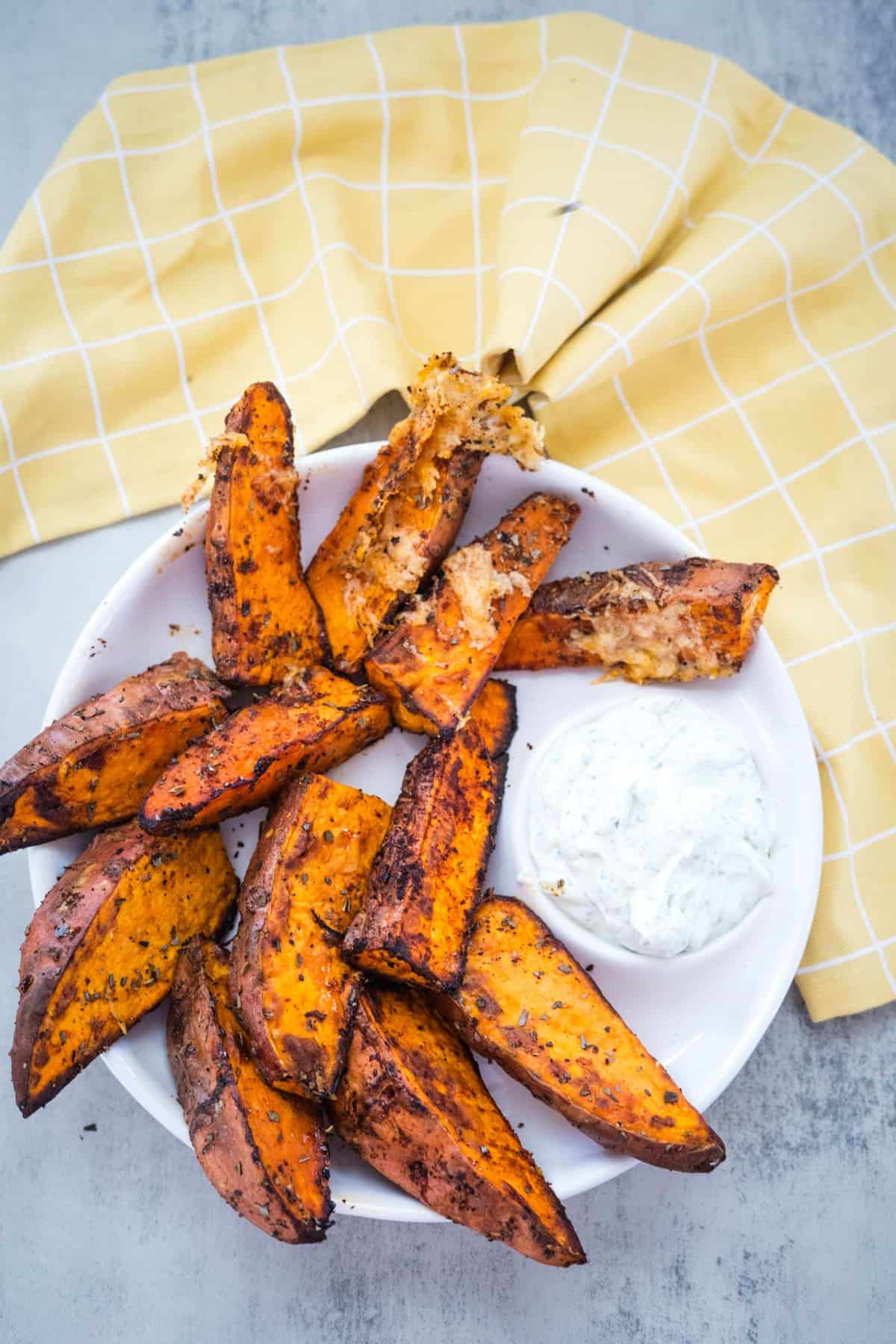 A plate of air fryer sweet potato wedges seasoned with herbs, served with a side of creamy dip. A yellow checkered cloth partially visible in the background.