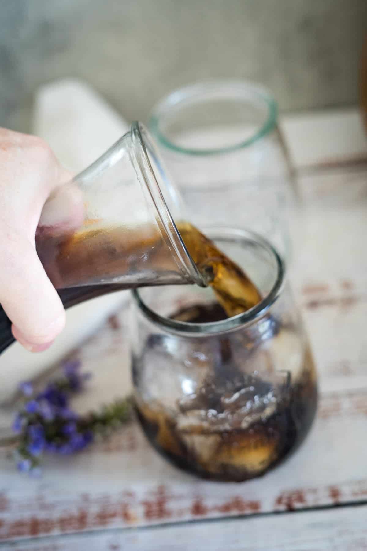 A hand is pouring cold brew coffee from a glass pitcher into a glass filled with ice cubes, with another empty glass, a towel, and lavender flowers in the background.