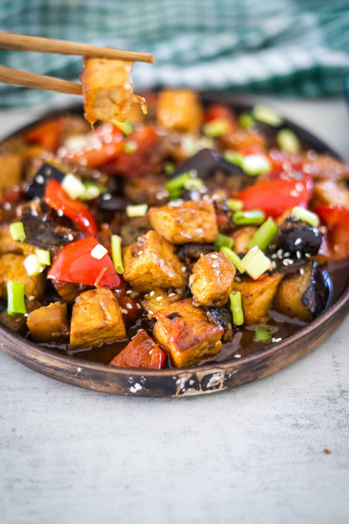 A plate of stir-fried tofu and vegetables, including red bell peppers, green onions, and tender eggplant, with chopsticks holding a piece of tofu.