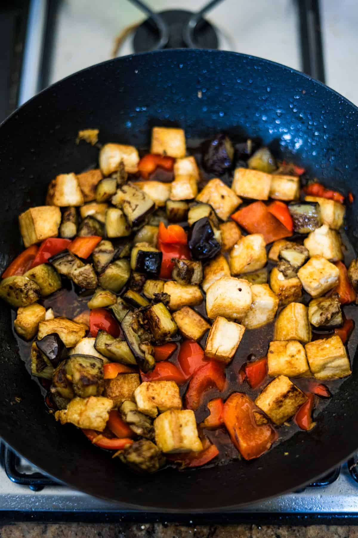 A frying pan on a stove sizzling with an eggplant tofu recipe, including diced tofu, vibrant red bell peppers, and succulent eggplant.