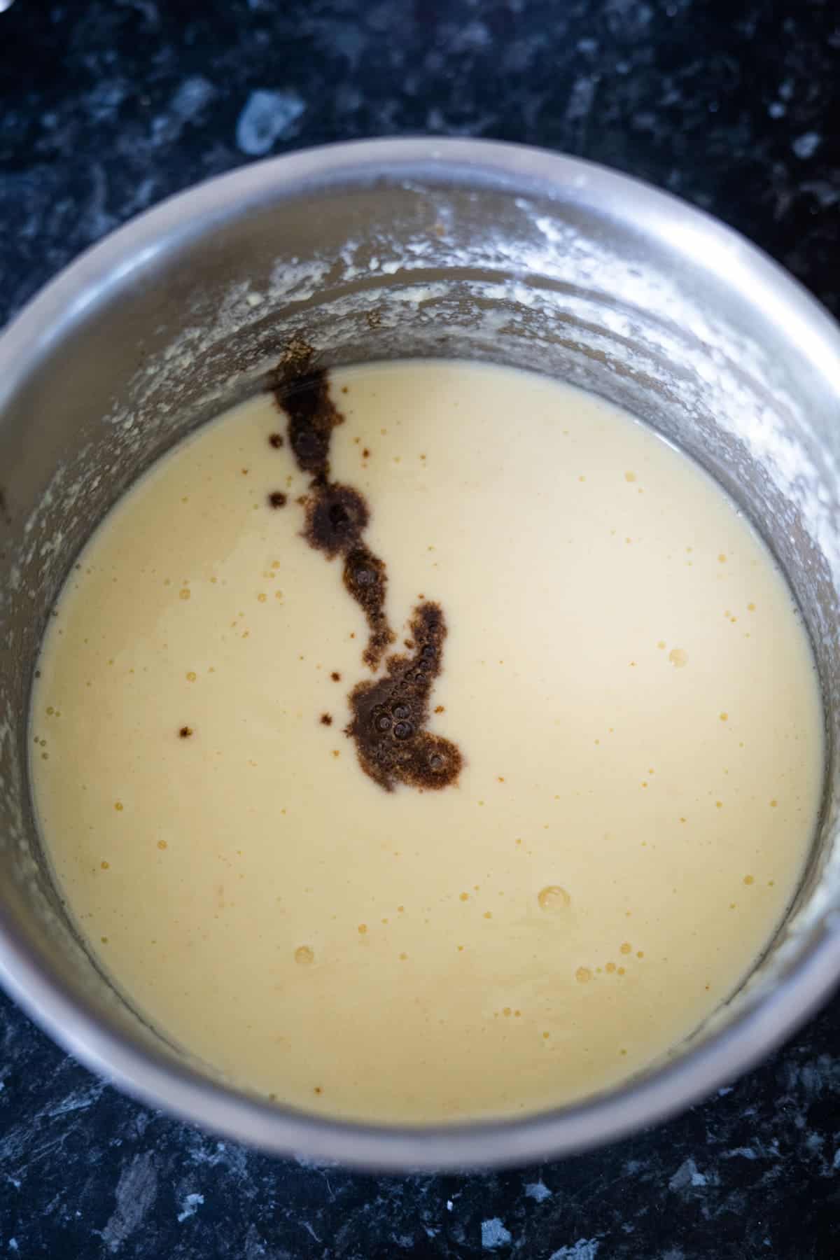 A metal bowl with a light yellow batter, resembling the base for keto vanilla ice cream, features a streak of brown liquid on the surface and sits on a dark countertop.