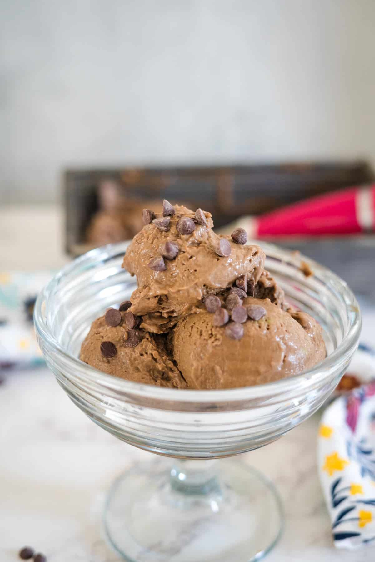 A glass bowl contains three scoops of decadent keto avocado ice cream topped with chocolate chips. The background includes a colorful napkin and an out-of-focus object.