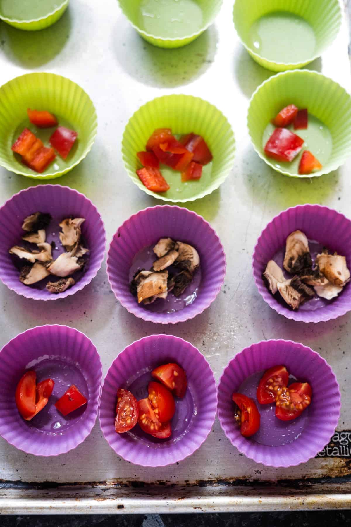 Silicone cupcake molds with assorted ingredients, including chopped cherry tomatoes, mushrooms, and red bell peppers, arranged on a baking sheet, perfect for making cottage cheese egg muffins.