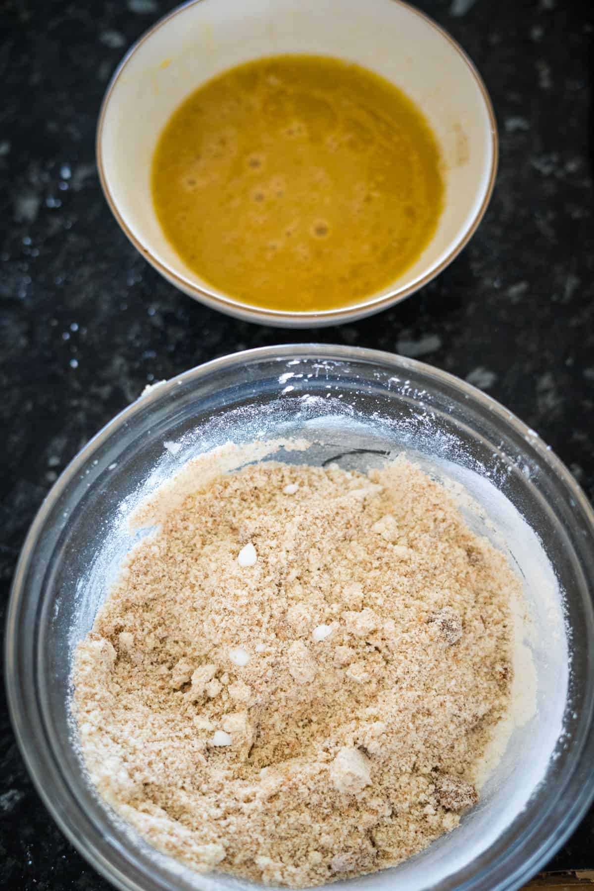 Two bowls on a dark countertop: one containing a whisked yellow mixture and the other filled with a crumbly flour blend.