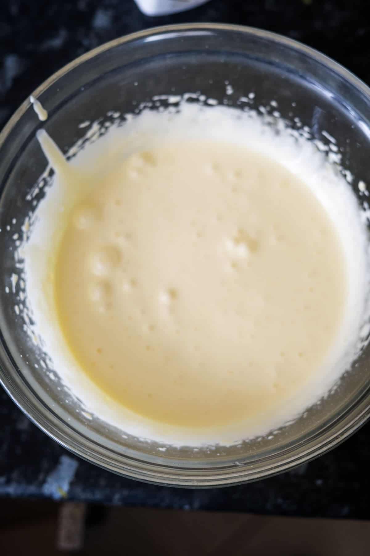 A clear glass bowl filled with a smooth, pale yellow keto vanilla ice cream mixture is placed on a dark surface. The creamy concoction appears slightly bubbly and rich in texture.