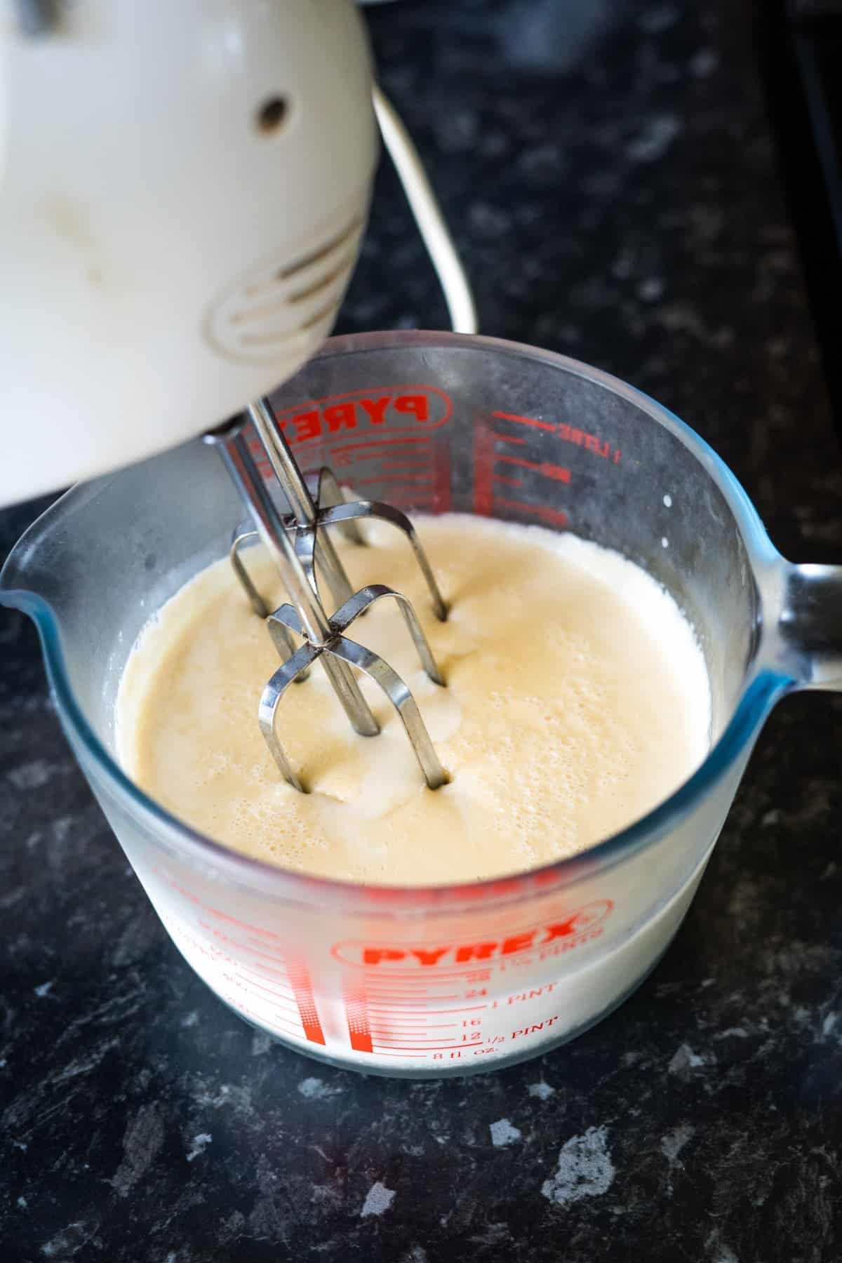 A hand-held electric mixer blending a creamy mixture for keto ice cream bars in a Pyrex measuring jug on a dark countertop.