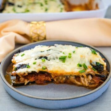 A slice of baked lasagna topped with melted cheese and chopped chives served on a grey plate, with a golden napkin ring and baking dish in the background.