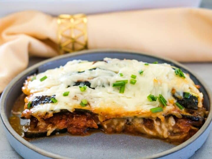 A slice of baked lasagna topped with melted cheese and chopped chives served on a grey plate, with a golden napkin ring and baking dish in the background.