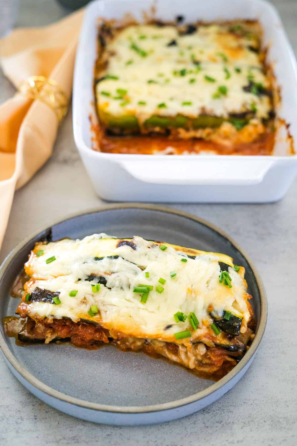 A slice of vegetable lasagna, reminiscent of zucchini parmigiana and garnished with chives, is served on a plate. A casserole dish with the remaining lasagna is in the background.