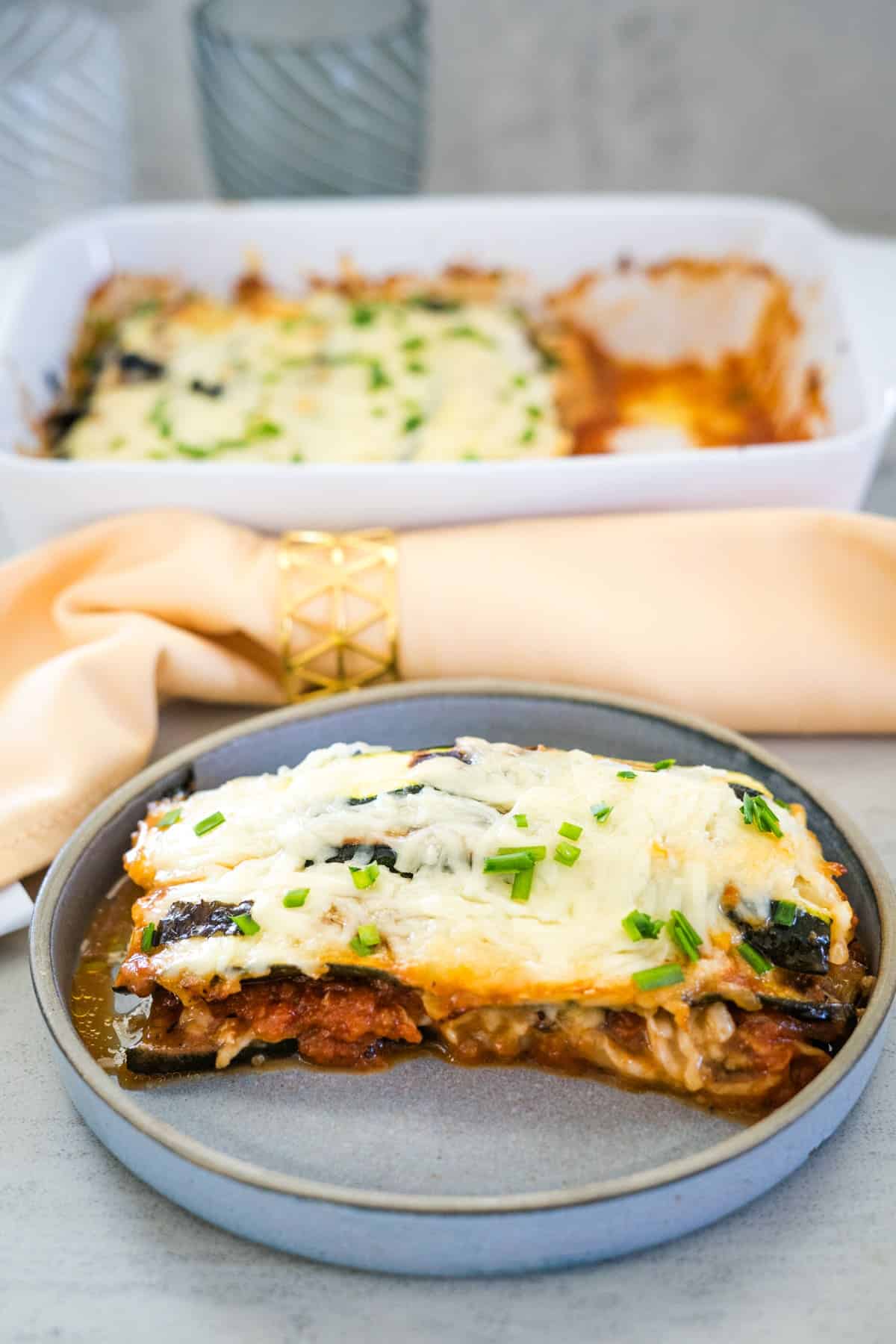 A plate with a slice of vegetable lasagna, reminiscent of zucchini parmigiana, topped with melted cheese and chives is in the foreground, with a baking dish containing more lasagna and a beige napkin in the background.