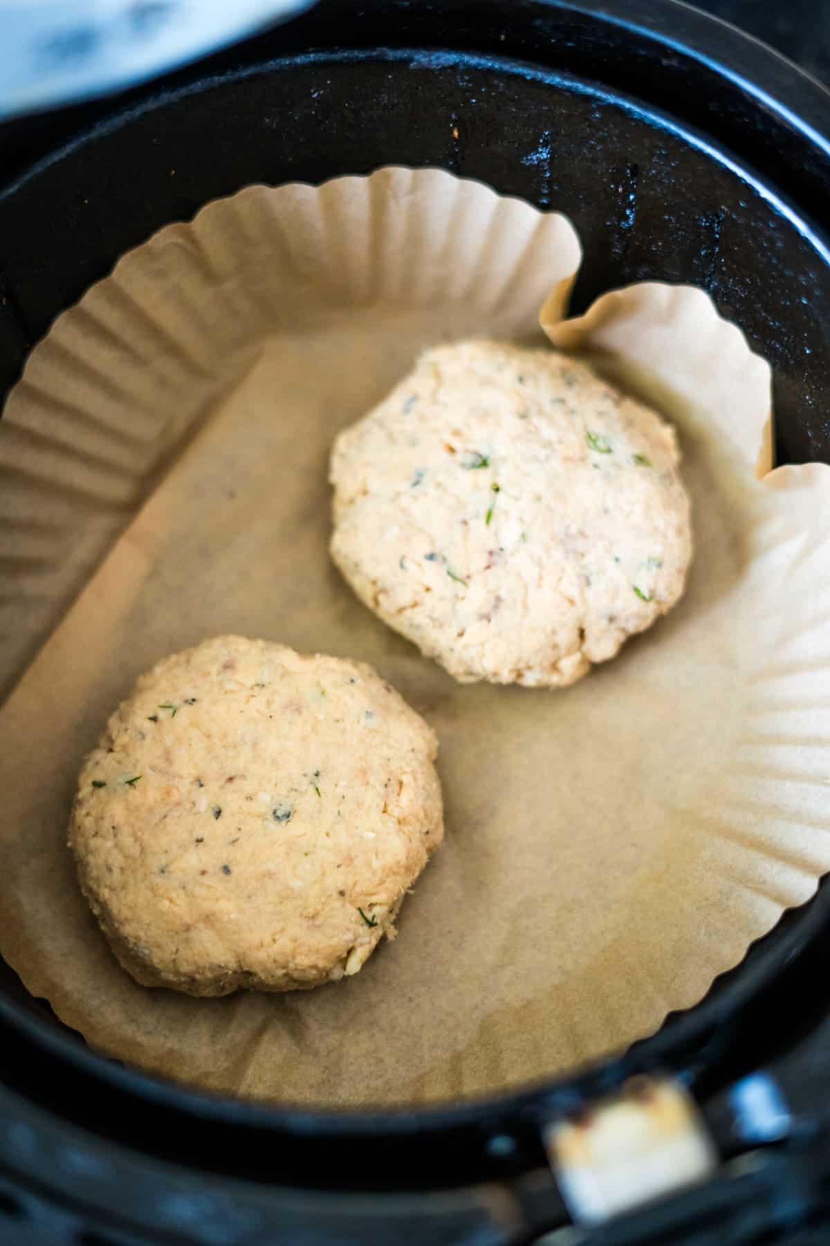 Two uncooked patties with herbs, placed on parchment paper inside a black air fryer.