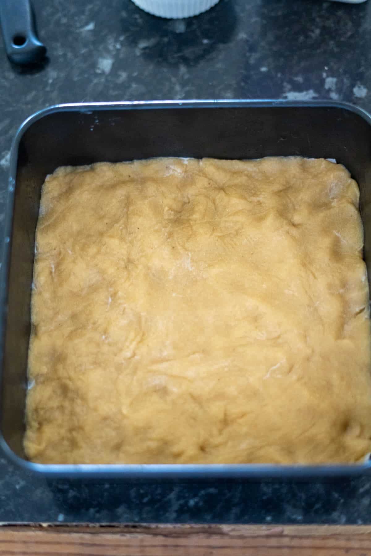 A square baking pan filled with unbaked dough sits on a dark countertop.