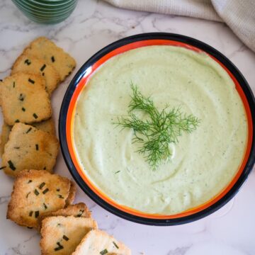 A bowl of creamy green dip garnished with dill, placed next to a stack of toasted bread slices sprinkled with herbs, on a marble surface.