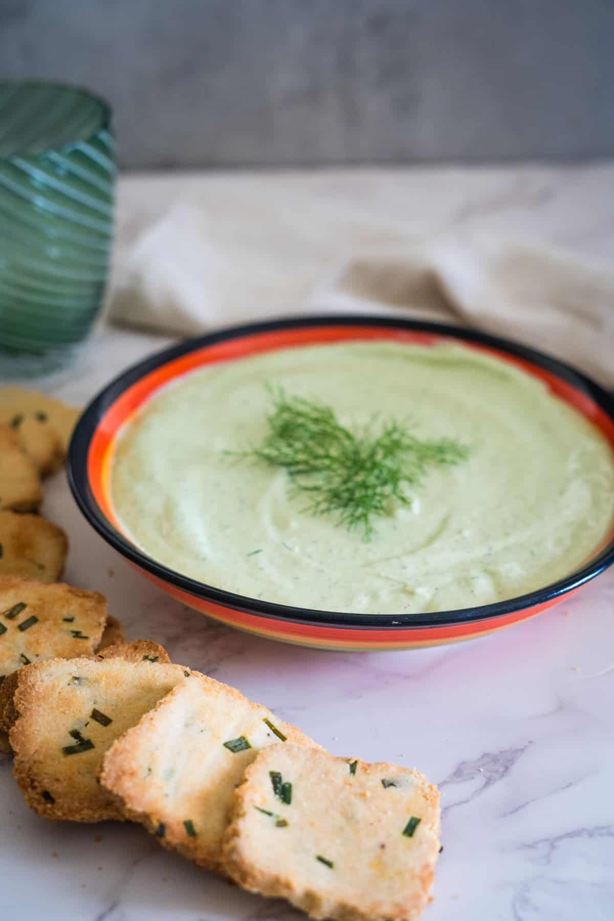 A bowl of creamy green cottage cheese avocado dip garnished with fresh herbs, surrounded by pieces of toasted bread sprinkled with chives, on a marble surface.