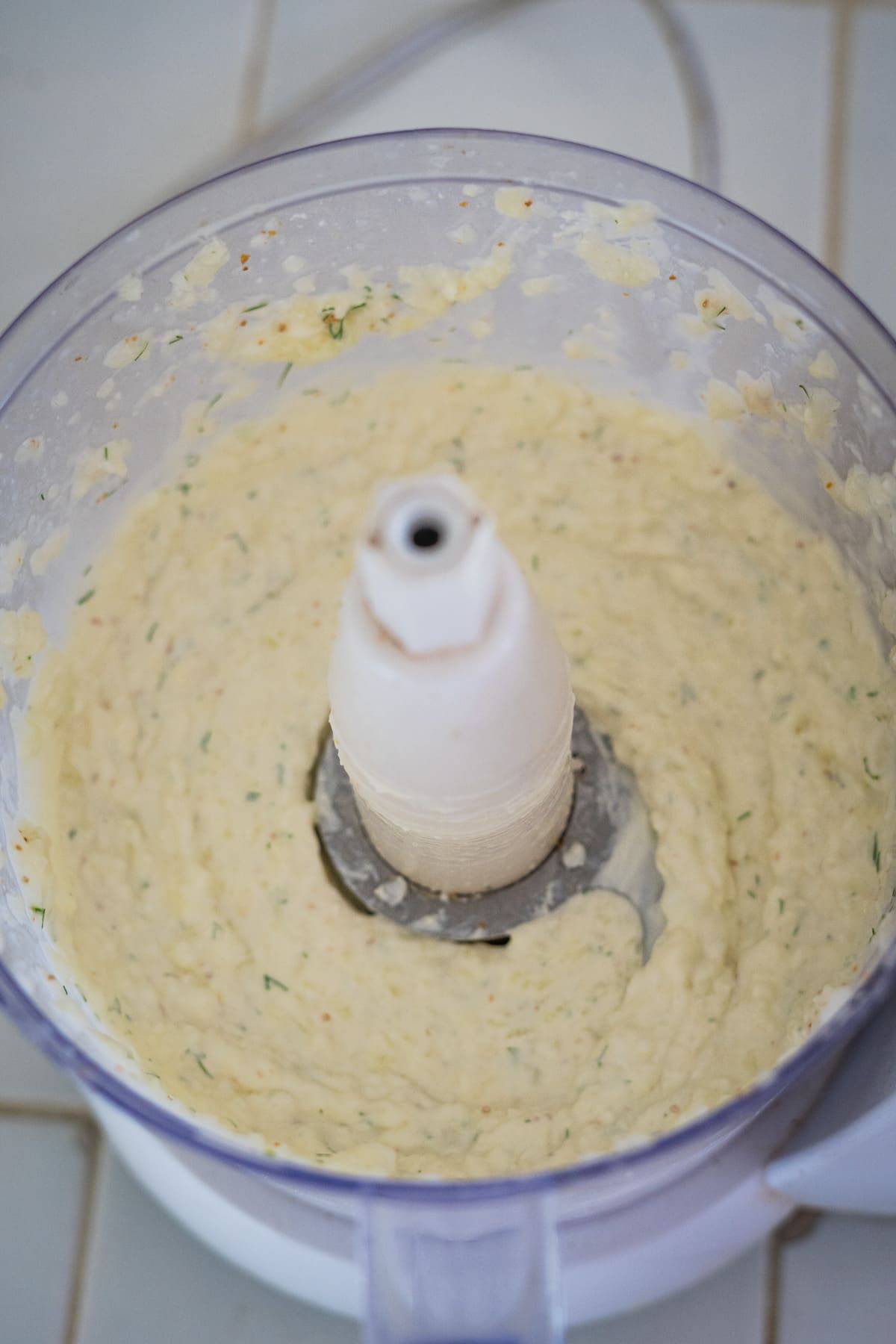 Top view of a food processor containing a creamy cucumber cream cheese dip with visible flecks of herbs, likely in the preparation of a blended dish.