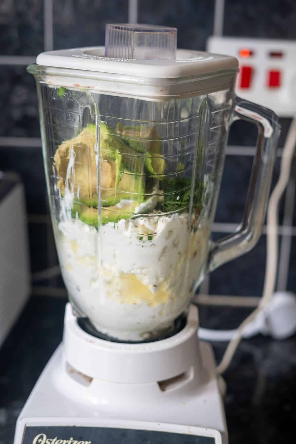 A blender containing ingredients such as avocado, cottage cheese, and other foods sits on a kitchen counter, ready to be blended.