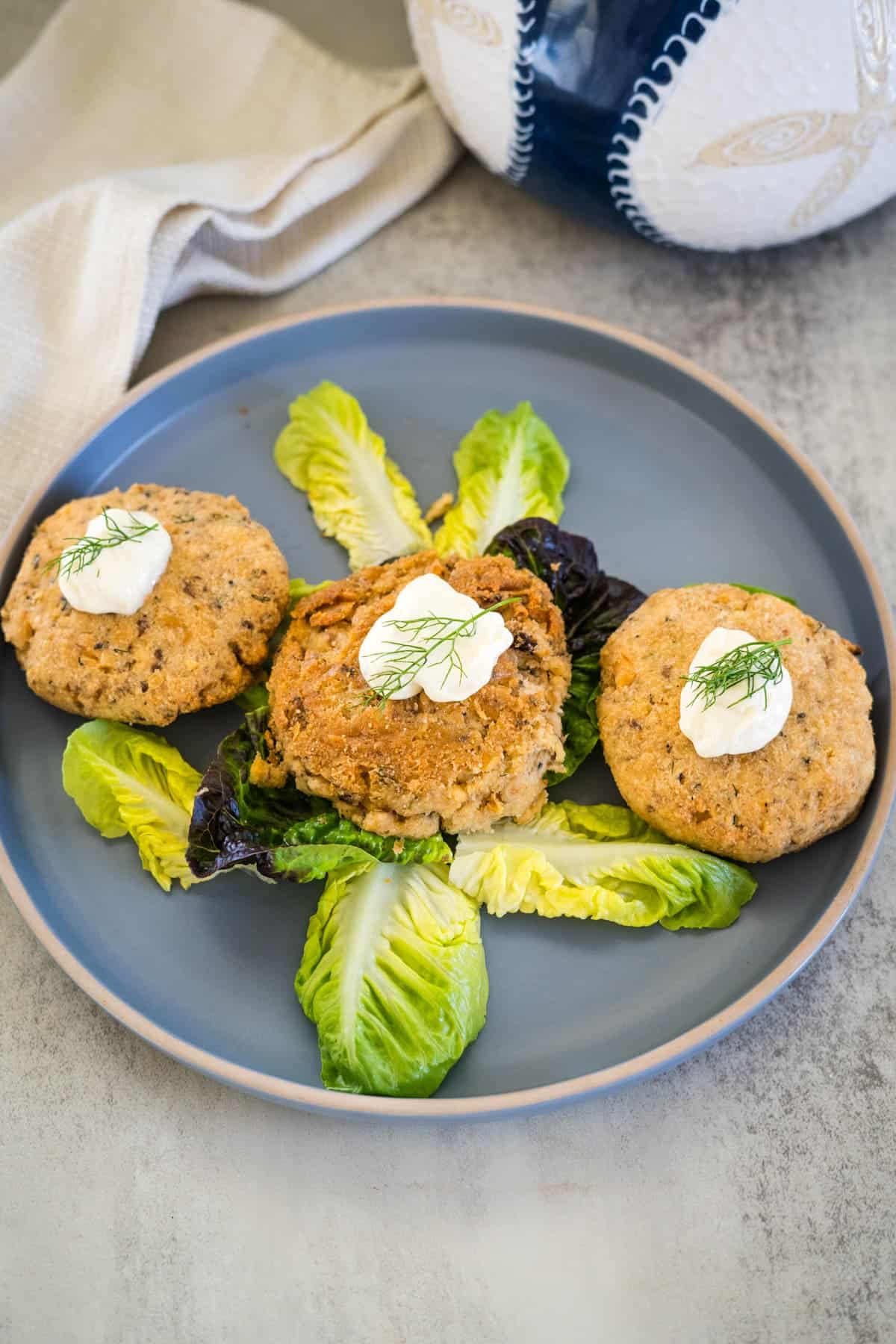 A blue plate with three salmon cakes garnished with dollops of white sauce and dill, arranged on top of green lettuce leaves. A beige napkin and part of a decorative bowl are visible in the background.