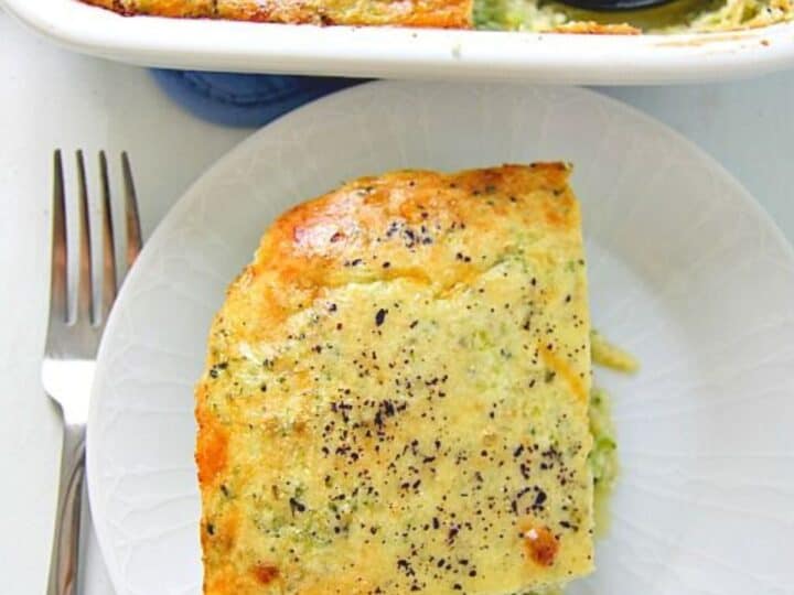 A slice of freshly baked savory pie with a golden, seasoned crust served on a white plate, next to a fork, with the baking dish and a serving spatula in the background.