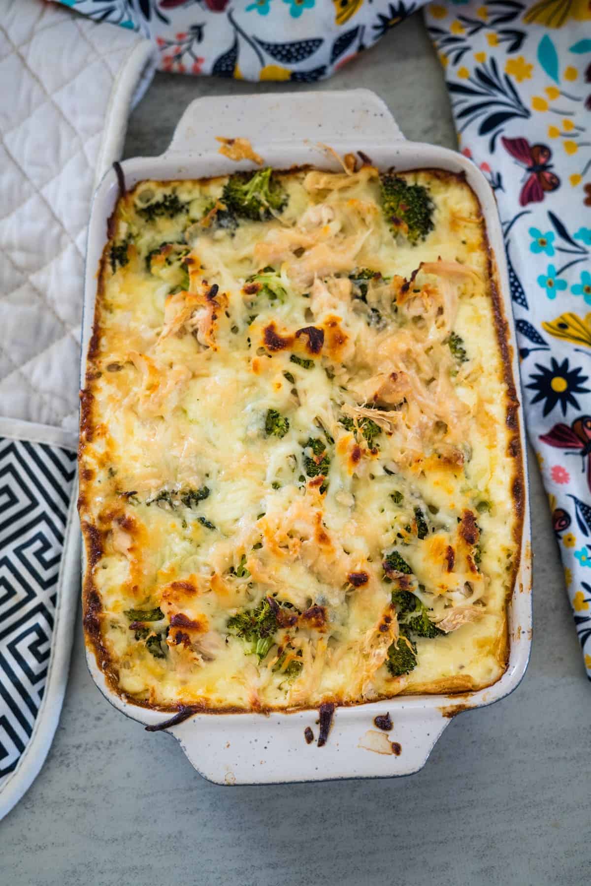 A baked casserole dish filled with a cheesy broccoli and chicken bake resting on a grey surface with patterned oven mitts beside it.