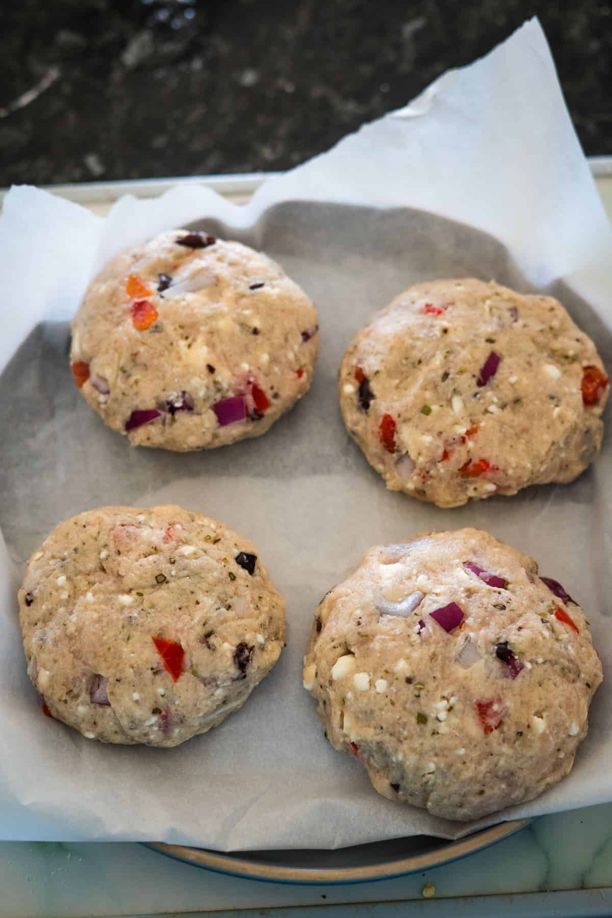 Four uncooked Greek chicken burgers made with visible chunks of vegetables and cheese placed on a sheet of parchment paper.