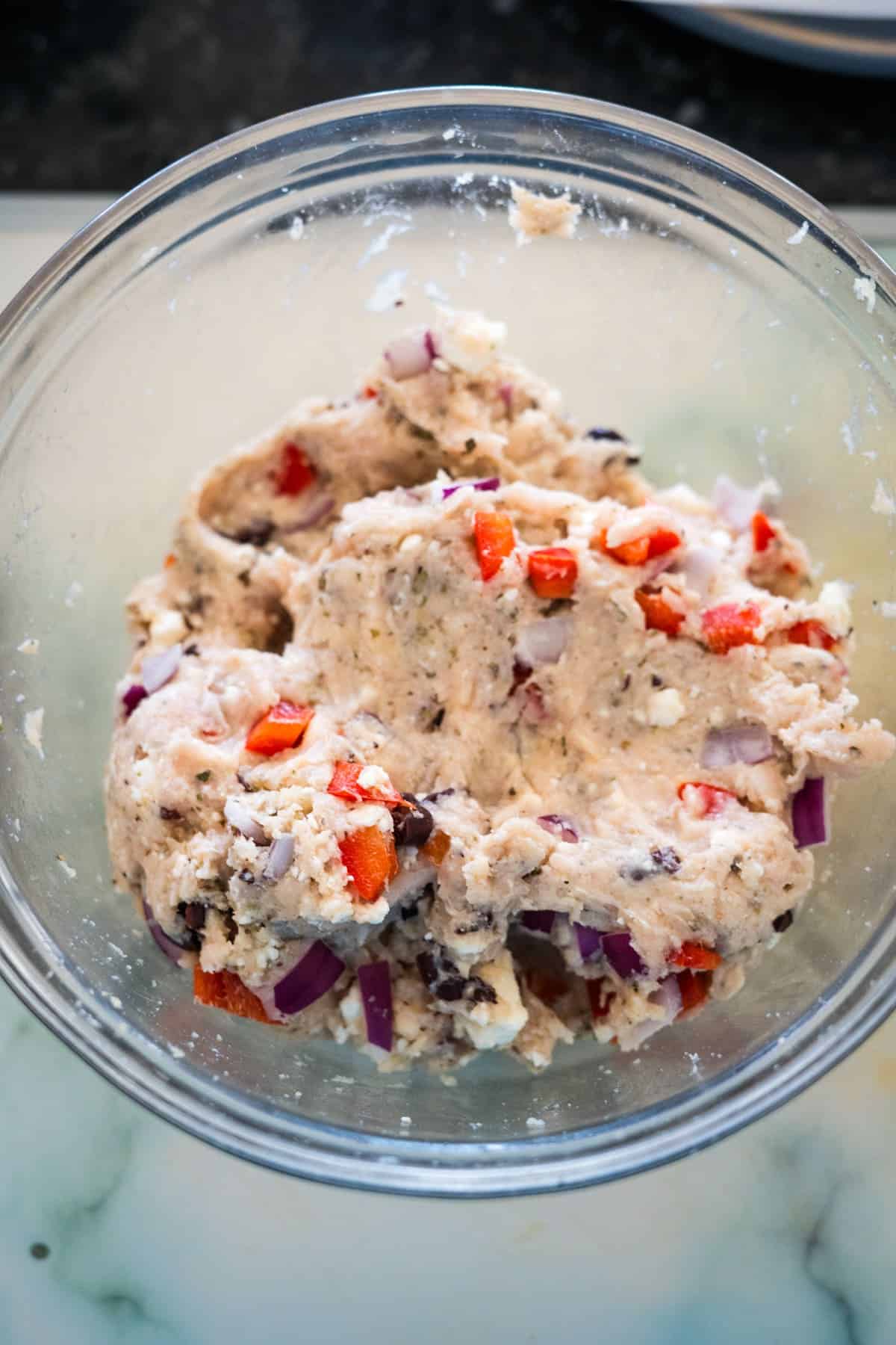 A glass bowl containing mixed dough for greek chicken burgers, with visible pieces of red bell peppers, red onions, and herbs on a light-colored surface.