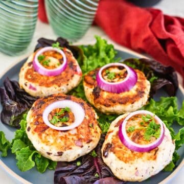 A plate with four grilled chicken patties, each garnished with a slice of red onion and a sprig of parsley, served on a bed of greens.