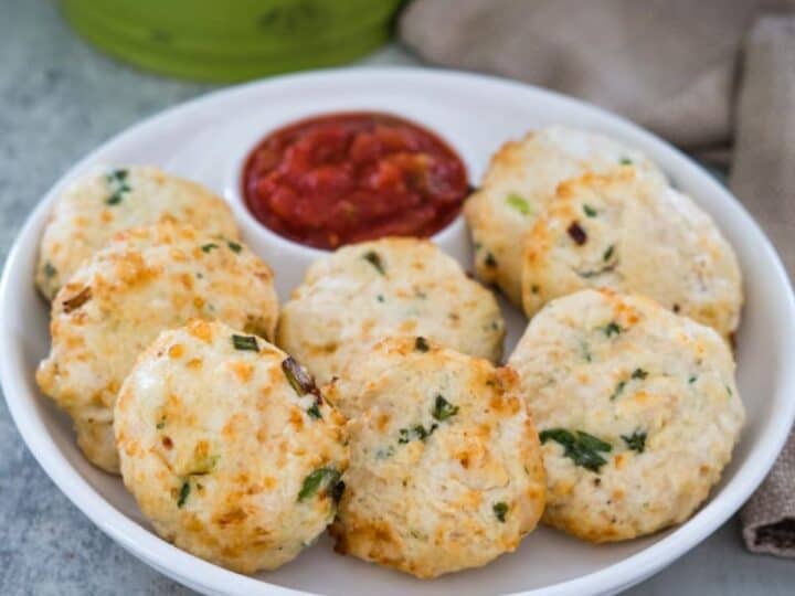 A plate of eight golden-brown savory biscuits garnished with herbs is served with a small bowl of red dipping sauce, placed on a table near a brown napkin.