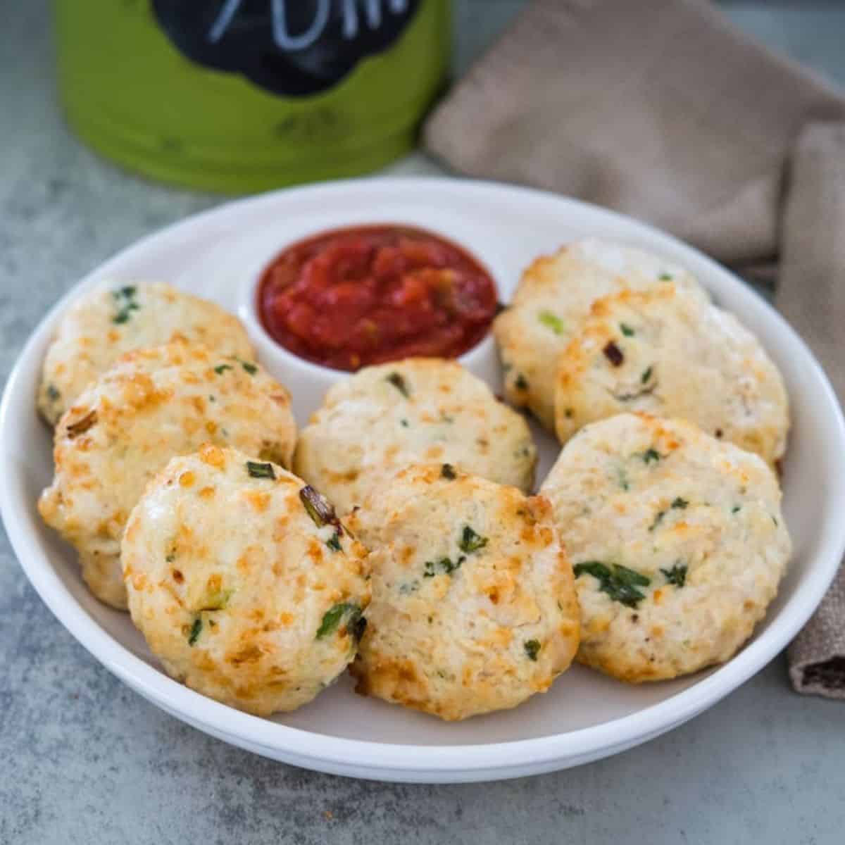 A plate of eight golden-brown savory biscuits garnished with herbs is served with a small bowl of red dipping sauce, placed on a table near a brown napkin.