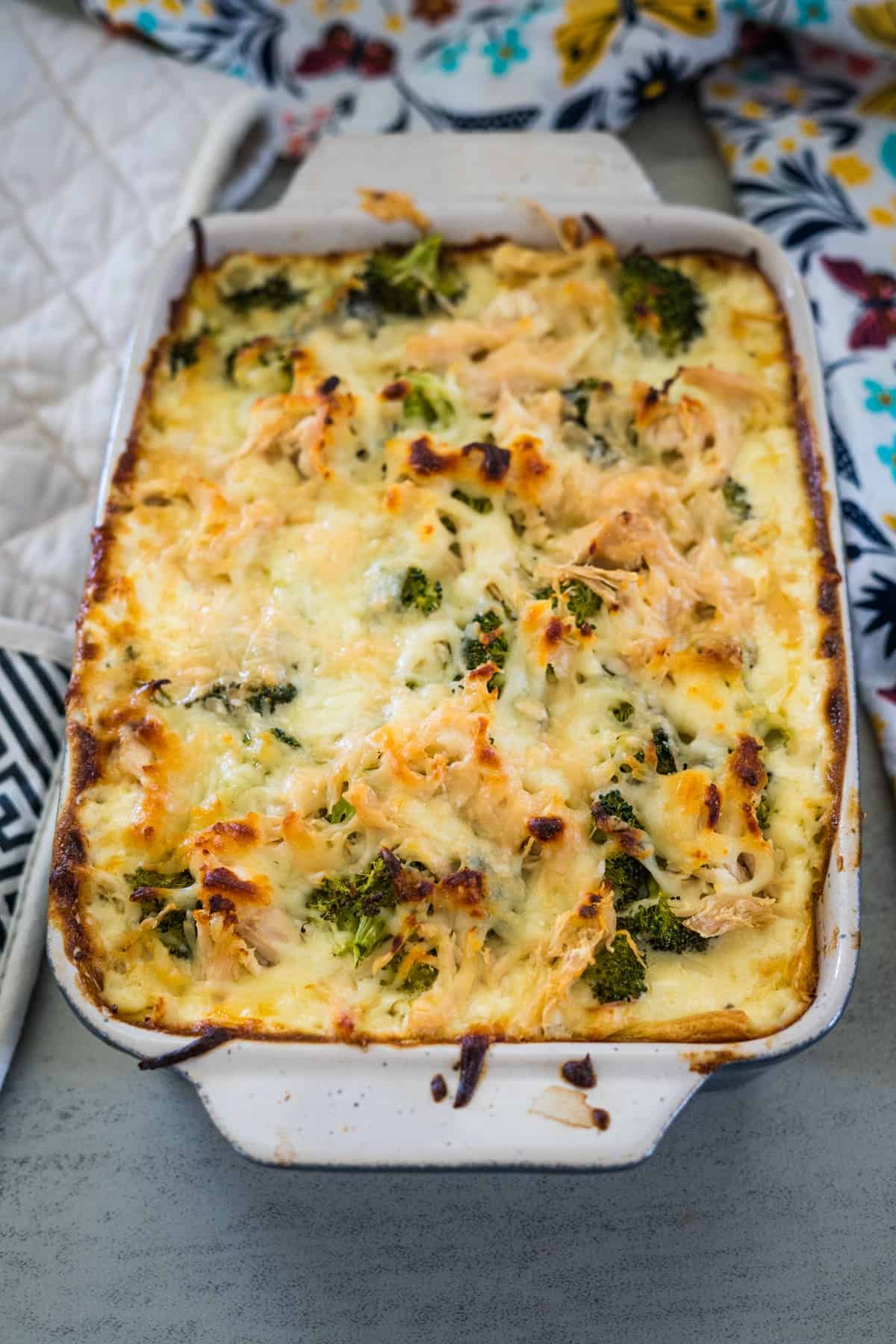 A baked casserole in a rectangular dish, topped with melted cheese, pieces of broccoli, and chicken, sits on a table with a patterned cloth in the background.