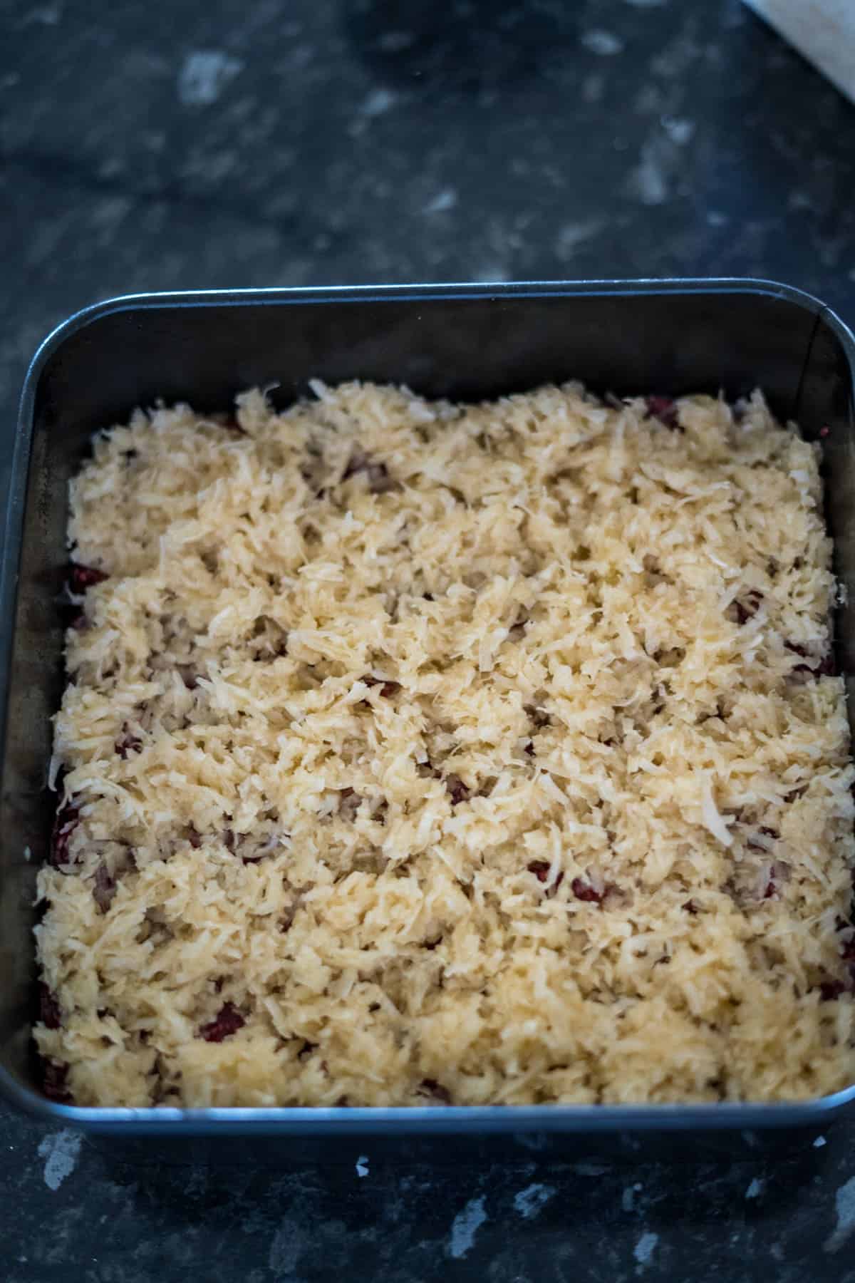 A square baking dish filled with an uncooked layer of shredded coconut mixture, placed on a dark countertop.