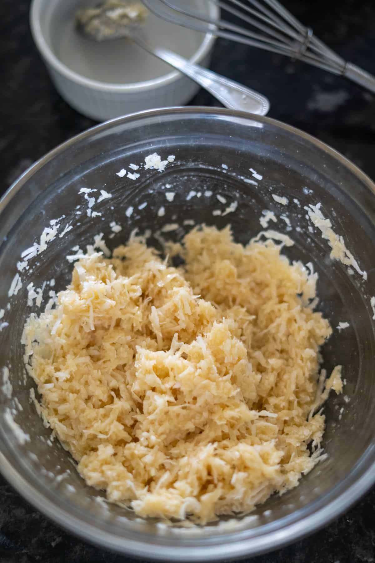 A glass bowl filled with a shredded mixture is placed on a dark countertop with a small white dish and a whisk in the background.