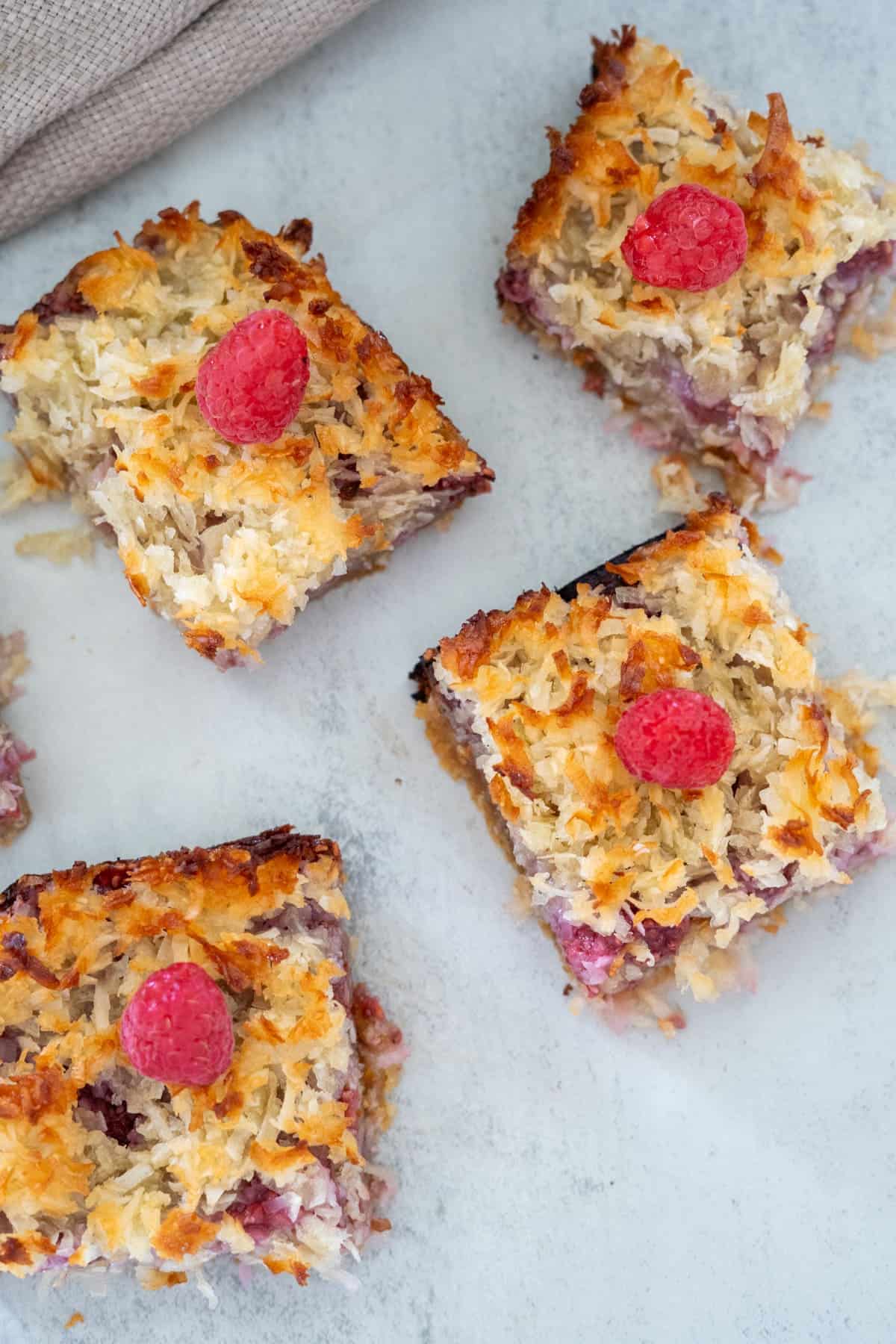 Four squares of coconut raspberry bars topped with raspberries are displayed on a light-colored surface.