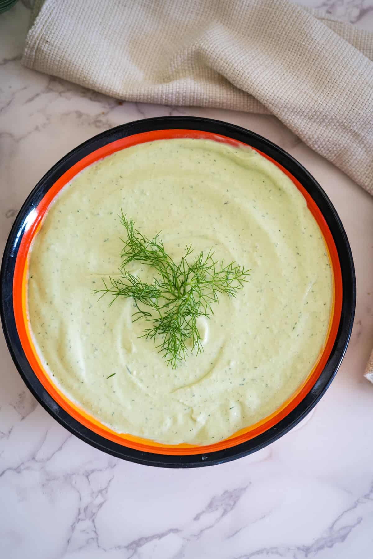 A bowl of creamy green soup, reminiscent of a rich avocado dip, garnished with fresh dill on a marble surface with a cloth napkin beside it.