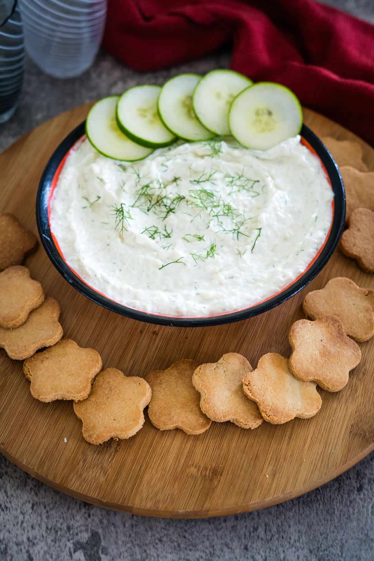 A bowl of dip garnished with dill and cucumber slices, surrounded by flower-shaped crackers on a wooden serving board.