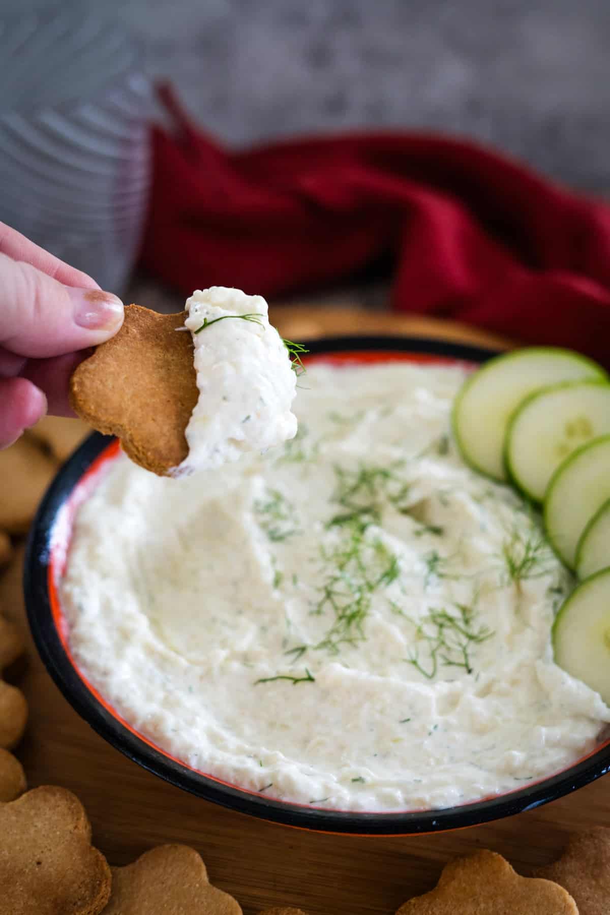 A hand is dipping a cracker into a bowl of cucumber cream cheese dip garnished with herbs, with cucumber slices and more crackers around the bowl.