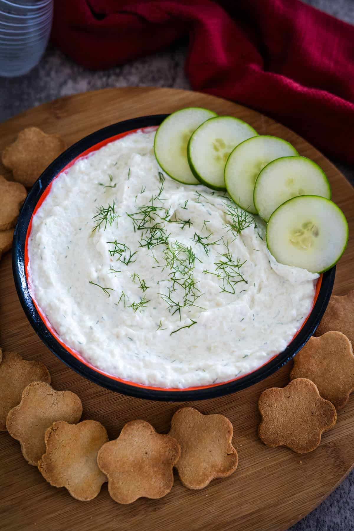 A bowl of creamy cucumber cream cheese dip garnished with fresh dill, accompanied by three cucumber slices and several flower-shaped crackers on a wooden surface. A red cloth is seen in the background.