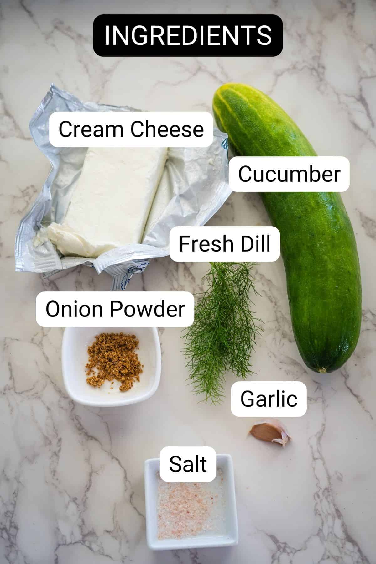 Ingredients for a recipe displayed on a marble surface: cream cheese, cucumber, fresh dill, onion powder, garlic clove, and salt.